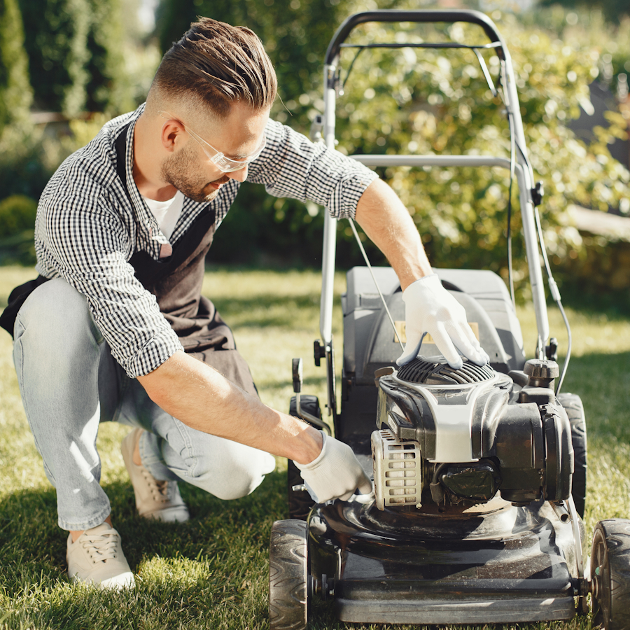 Man outside kneeling by a push lawn mower about to start it